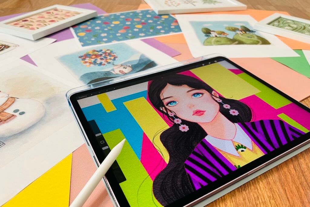 photo-4 An iPad showing a girl with long hair wearing magenta and black colored clothes, standing in front of vibrant background. A number of drawings on paper and in frames artistically arranged around the iPad.