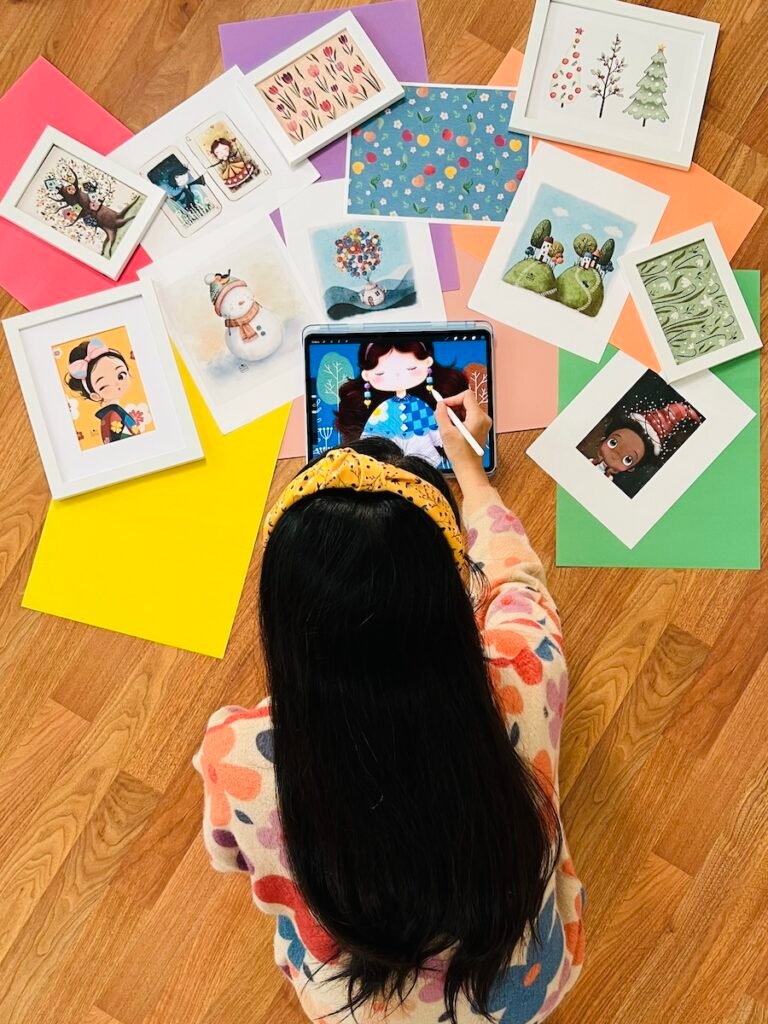 photo-5 An artist sitting on a wooden floor, drawing a cute whimsical girl on ipad. The artists work are artistically placed on the floor around her.